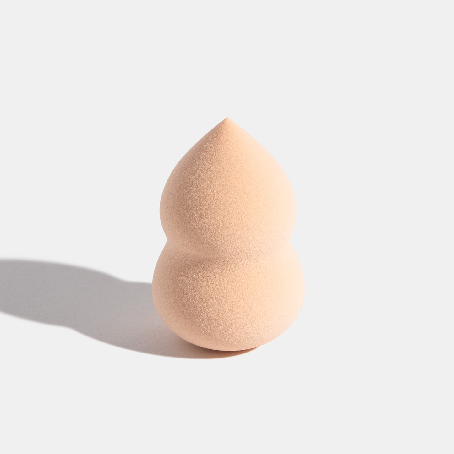 A tan makeup sponge standing upright on a white surface, casting a soft shadow to the left.