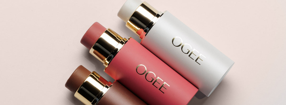 Three uncapped Ogee face sticks in white, coral pink, and deep brown, each with a gold band, lie diagonally on a blush pink surface.