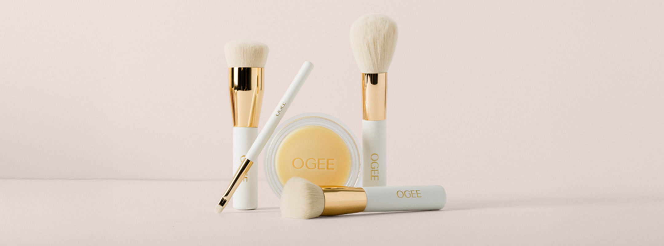 A collection of four white and gold Ogee makeup brushes arranged around a clear round container of Ogee product against a light pink background.