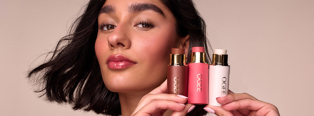 A woman with dark, tousled hair and rosy cheeks smiles softly while holding Ogee’s Crystal Contour Collection up beside her face.