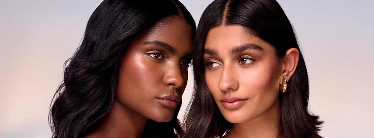 Two people with long dark hair pose cheek to cheek against a soft pink and lavender backdrop, both wearing glowing skin, defined brows, and soft nude lips, with one wearing a gold teardrop earring.