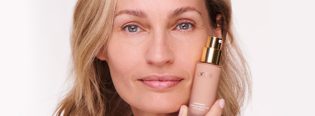 A woman with tousled blonde hair and a natural, lightly freckled complexion holds a frosted glass Ogee foundation bottle with a gold cap against her cheek.