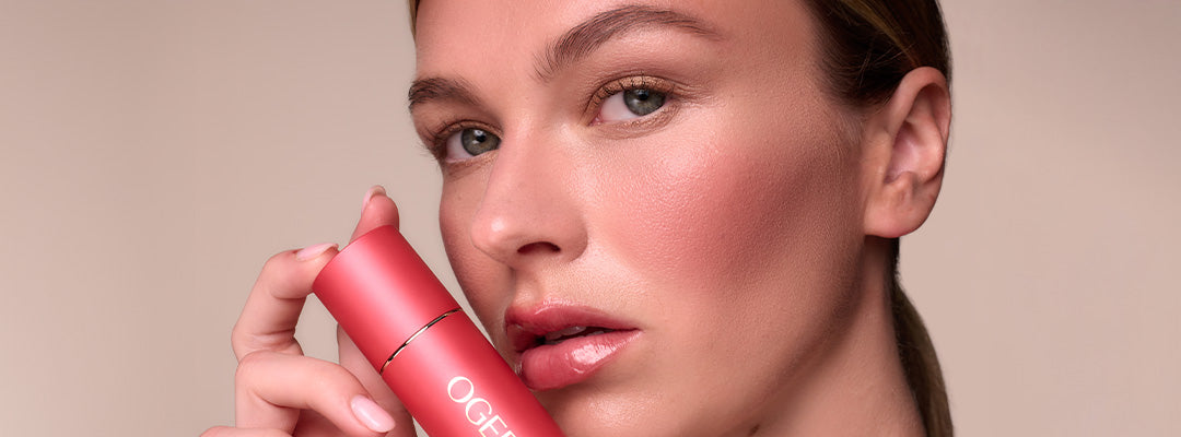 A woman holds an Ogee cream blush stick up to her cheek, which shows a natural rosy flush.