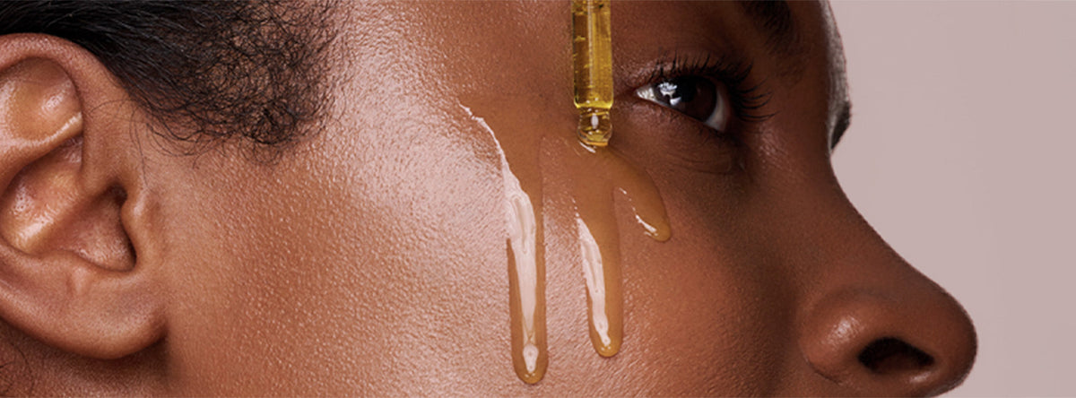 A close-up side profile of a person with deep brown skin as a glass dropper releases golden oil that drips down their cheek.