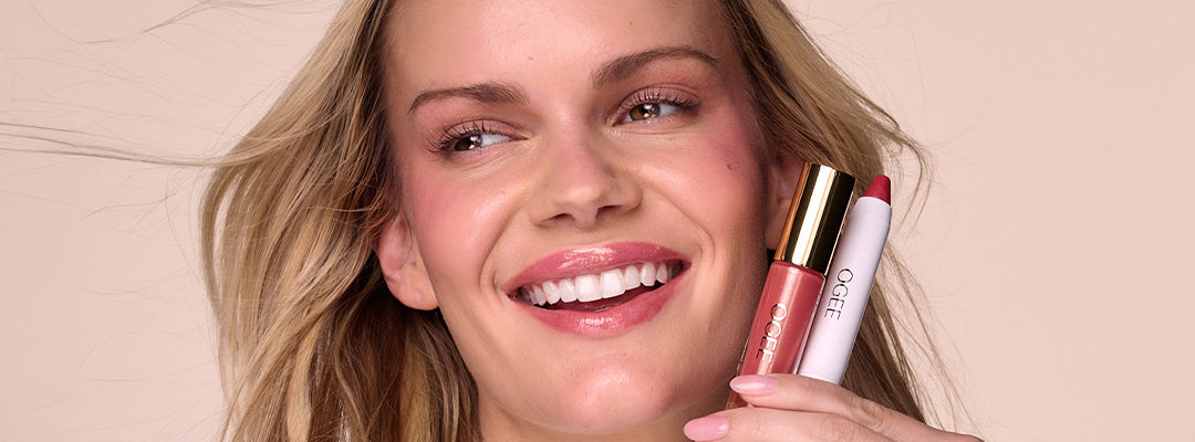 A smiling woman with wind-blown hair holds an Ogee lip gloss and lip oil up beside her face.