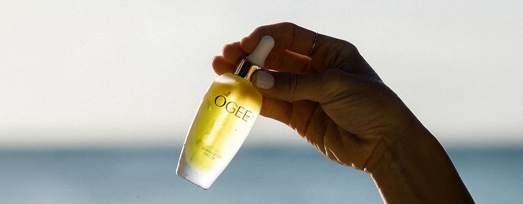 “A close-up shot of a hand holding a bottle of Ogee Jojoba Glow Face Oil against a blurred background.”