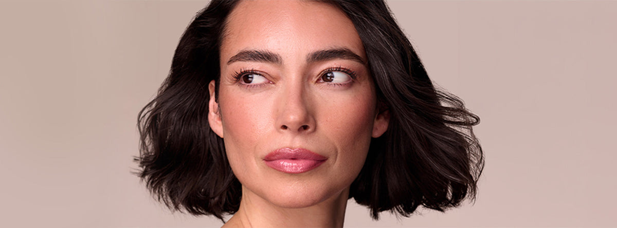A person with a dark, shoulder-length bob glances to the side against a muted pink backdrop, wearing soft blush, groomed brows, and a glossy pink lip.