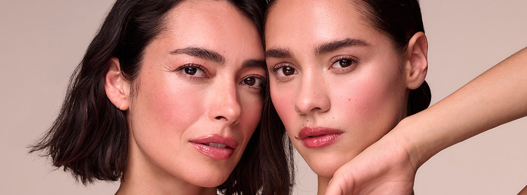 Two women with dark hair pose cheek to cheek against a soft pink background, both with dewy, hydrated-looking skin, rosy cheeks, and natural lips.