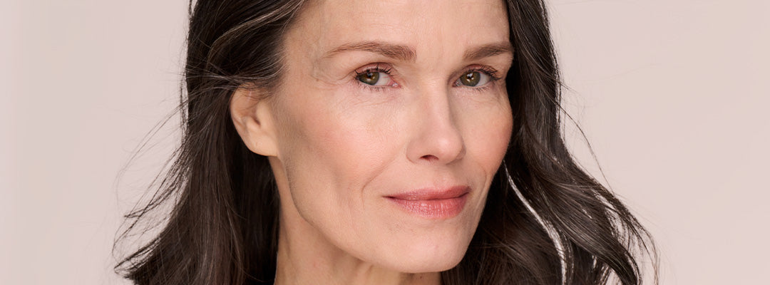 A mature woman with long, dark hair streaked with gray smiles softly at the camera, showing smooth, radiant skin and a natural makeup look against a pale pink background.