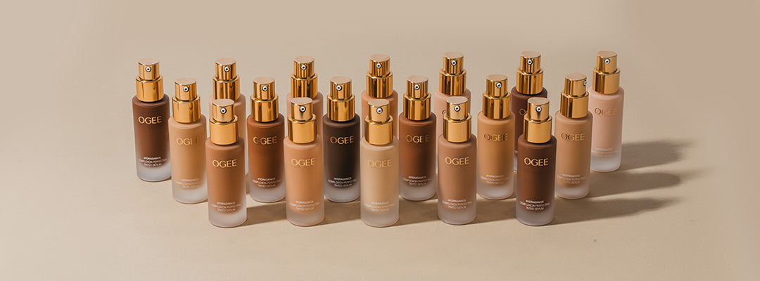 A staggered lineup of frosted glass Ogee Complexion Perfecting Serum Foundation bottles with gold pump tops on a beige surface, showing a wide range of shades from fair to deep.