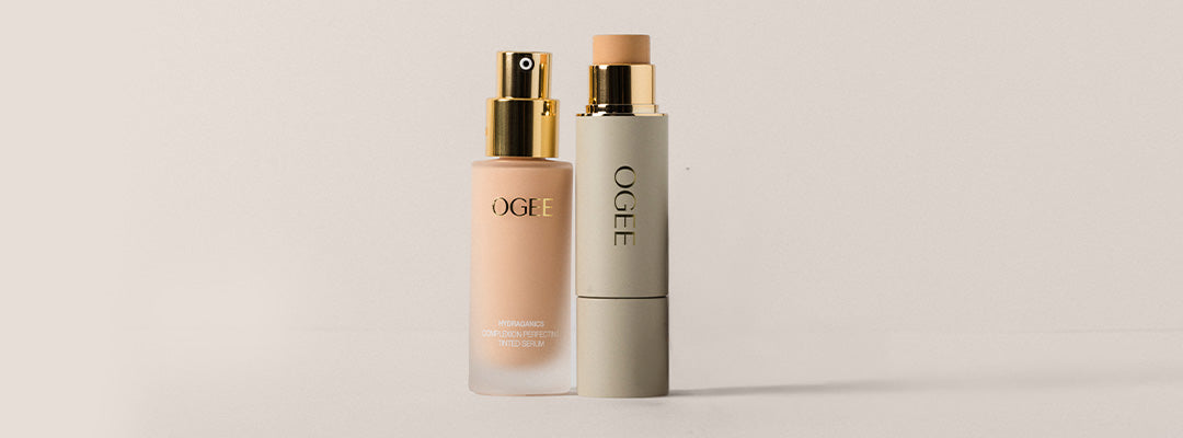 A frosted glass Ogee Complexion Perfecting Serum Foundation bottle with a gold pump top stands beside a taller sage green Ogee Sculpted Complexion Foundation Stick with its cap removed, set against a soft neutral background.