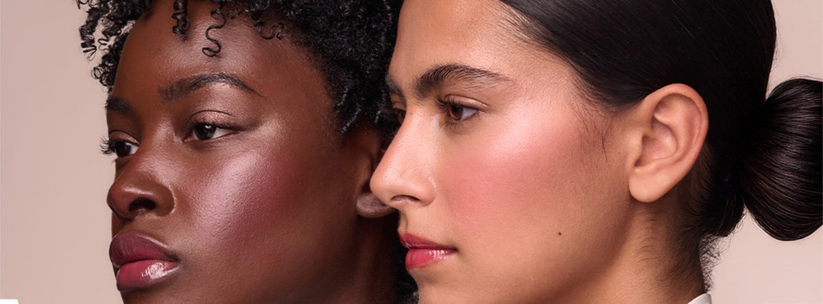 Two people pose cheek to cheek in profile against a dusty pink backdrop, one with curly dark hair and glossy plum lips and the other with dark hair in a low bun, flushed pink cheeks, and red lips.