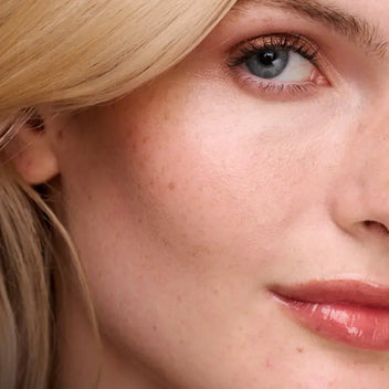 A close-up of a person with blonde hair and freckles, wearing shimmering copper eyeshadow and a glossy lip.