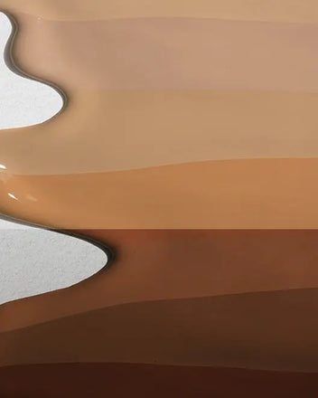 A top-down, close-up shot shows several thick, glossy streaks of liquid foundation in a range of shades. 