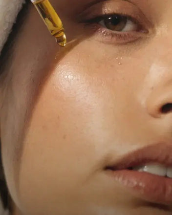 A close-up shot of a person's face shows them applying a drop of facial oil from a glass dropper onto their cheek. 