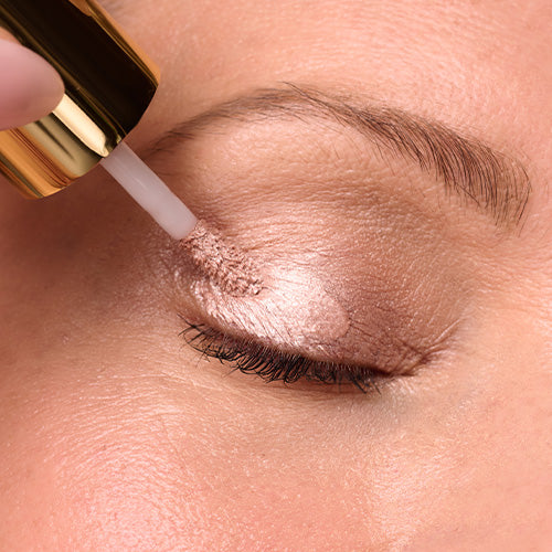 A close-up of a person’s closed eye as a gold-capped applicator glides a shimmery, pale pink liquid eyeshadow across their eyelid.
