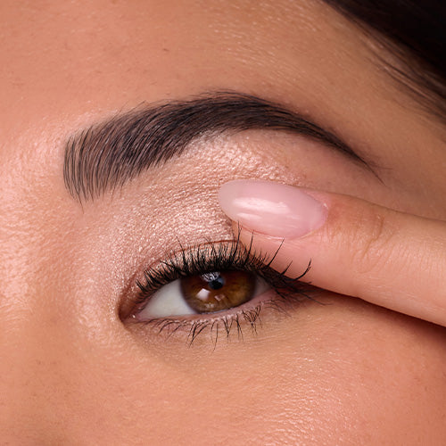 A close-up of a person’s brown eye with defined brows and long lashes as they blend a shimmery champagne eyeshadow onto their lid with their fingertip.