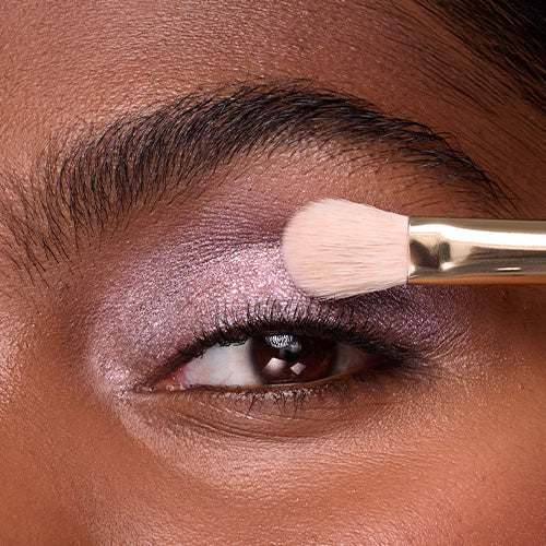 A close-up of a person’s eye with deep brown skin as a fluffy makeup brush with a gold ferrule sweeps a shimmery lavender eyeshadow across their lid.