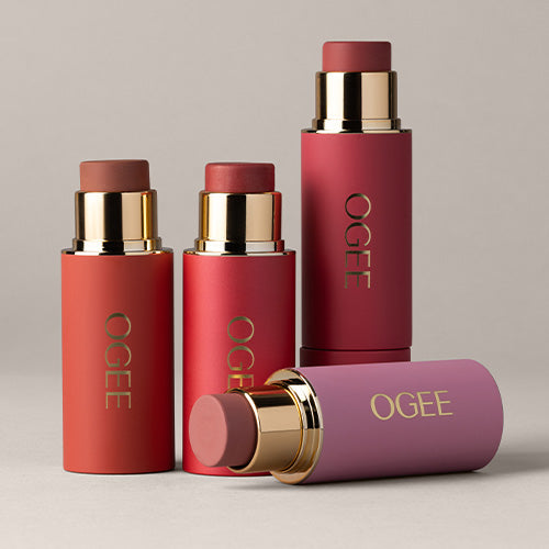 Three Ogee blush sticks with gold bands stand on a neutral surface, with a fourth stick lying on its side in front.