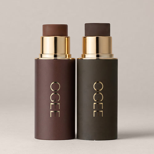 Two uncapped Ogee Sculpted Face Sticks with gold bands stand side by side on a neutral background, one in a deep espresso shade and the other in a dark olive shade.