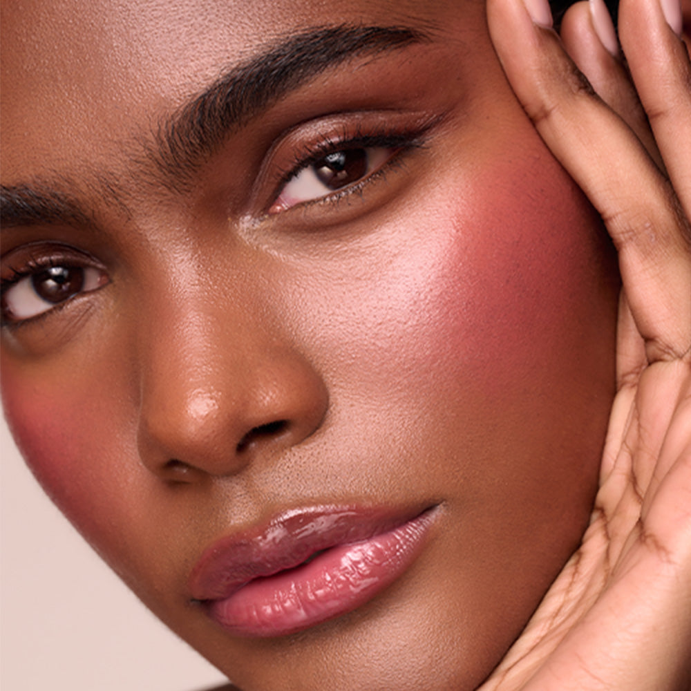 A close-up of a person with deep brown skin resting their fingertips on their temple, wearing berry-toned blush on the cheeks and glossy pink lips.