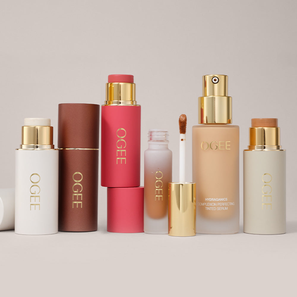 A lineup of Ogee makeup products with gold caps on a pale pink surface, including face sticks in white, brown, and coral, a liquid applicator, a tinted serum pump bottle, and a light beige stick.