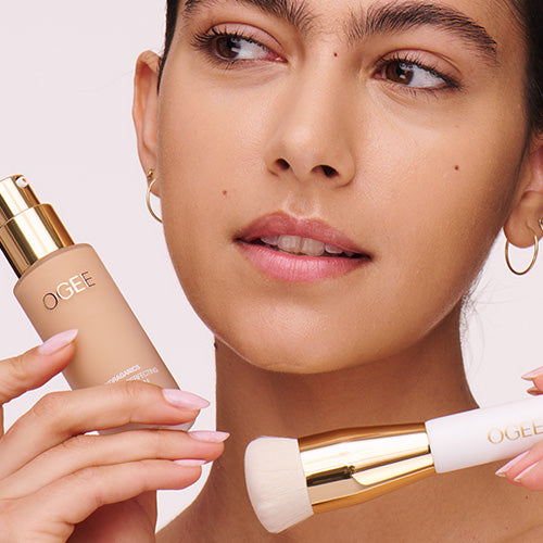 A woman with dark hair pulled back and gold hoop earrings holds an Ogee Complexion Perfecting Serum Foundation bottle and a gold-handled foundation brush near her face, looking off to the side.