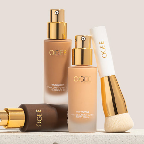 Two frosted glass Ogee Complexion Perfecting Serum Foundation bottles with gold pump tops stand beside a gold-handled foundation brush, with a deeper-shade bottle lying on its side in front.