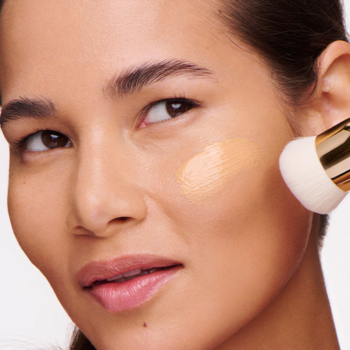 A close-up of a woman with dark hair and a smooth, even complexion as a gold-handled foundation brush buffs a sheer beige foundation onto her cheek.