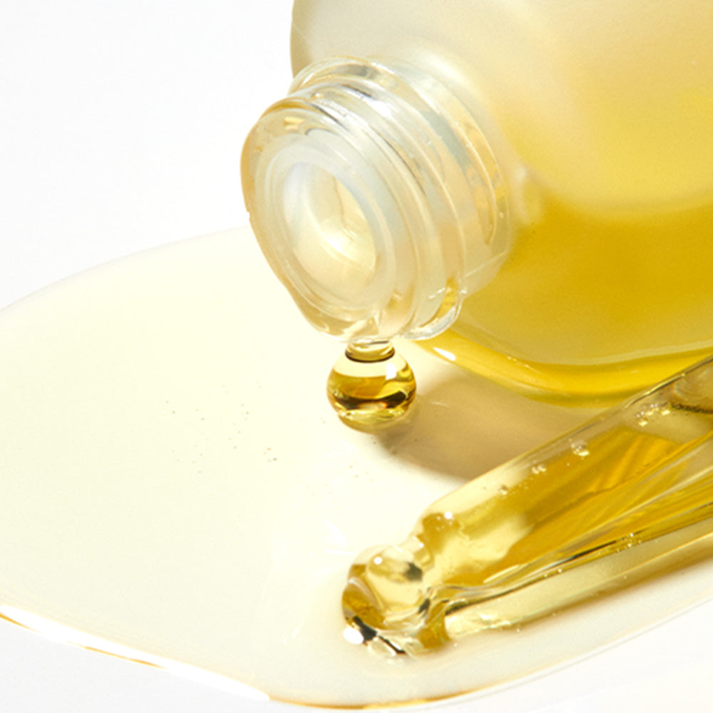 A frosted glass bottle tipped on its side spills golden oil onto a white surface beside a glass dropper filled with oil.