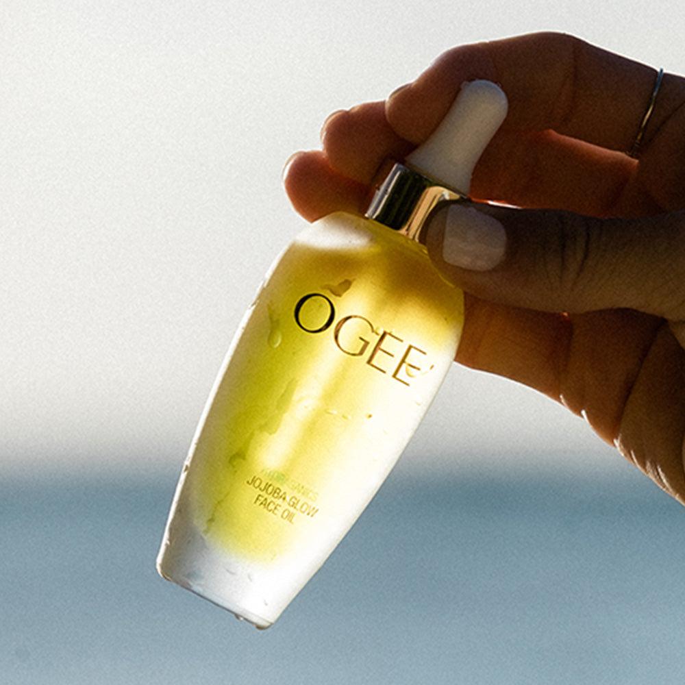 A hand holds up a frosted bottle of Jojoba Glow Face Oil, with a white dropper cap and gold collar, against a blurred background of sky and water.