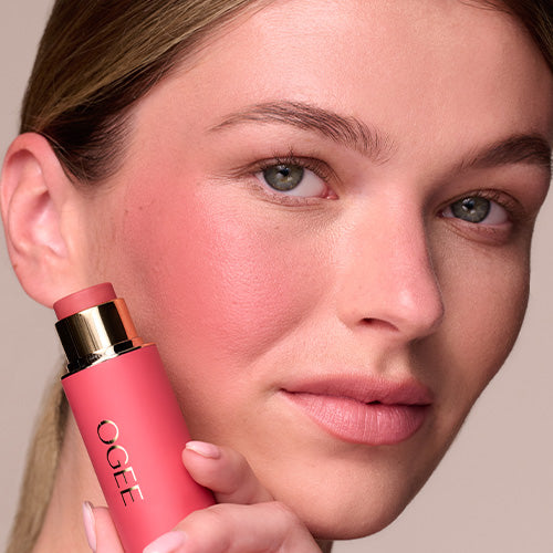 A woman with light brown hair and rosy, flushed cheeks holds a coral pink Ogee Sculpted Face Stick with a gold cap against her face, looking at the camera.