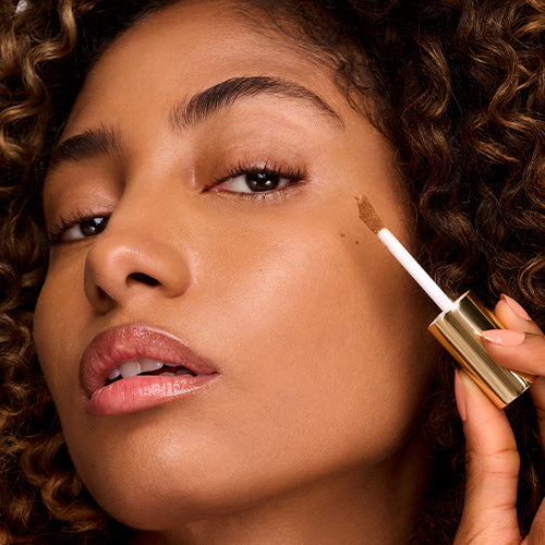 A woman with curly dark hair and a glowing complexion applies a liquid product to her cheekbone with a doe-foot wand, holding a gold-capped Ogee tube.