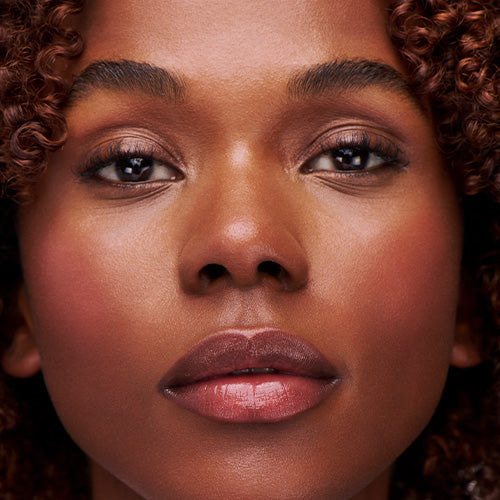 A close-up portrait of a woman with deep brown skin and curly hair, wearing a warm rosy blush and glossy lips as she looks directly at the camera.