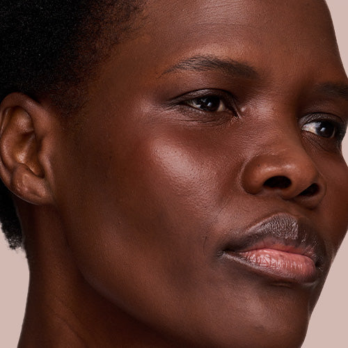 A close-up portrait of a woman with deep brown skin and curly hair, wearing a warm rosy blush and glossy lips as she looks directly at the camera.