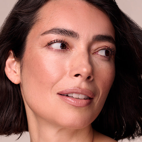 A close-up of a mature woman with dark hair, defined brows, and luminous skin with a hint of rosy blush, glancing off to the side over her shoulder.