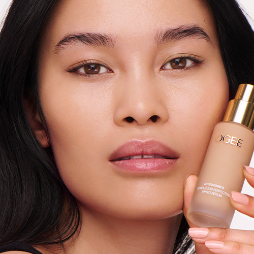A woman with dark hair pulled back and glowing, even-toned skin holds a frosted glass Ogee Complexion Perfecting Serum Foundation bottle up beside her cheek.