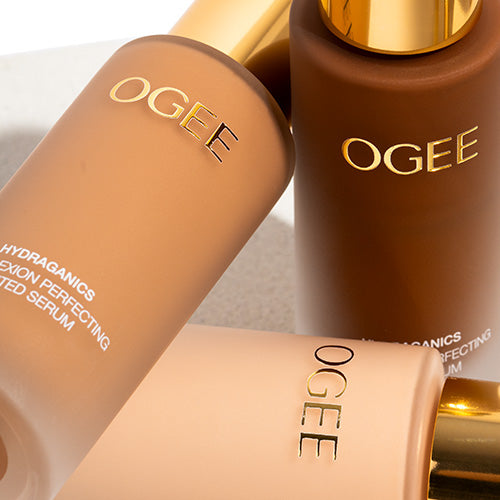 A close-up of three Ogee Complexion Perfecting Serum Foundation bottles with gold caps lying at angles, showing light, medium, and deep brown shades with the Ogee name printed on each.