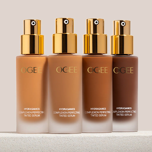 Four frosted glass Ogee Complexion Perfecting Serum Foundation bottles with gold pump tops stand in a row on a white surface, filled with shades ranging from light tan to deep brown.