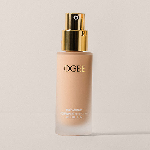 A single frosted glass bottle of Ogee Complexion Perfecting Serum Foundation with a gold pump top, filled with a light beige formula, stands against a soft neutral background.