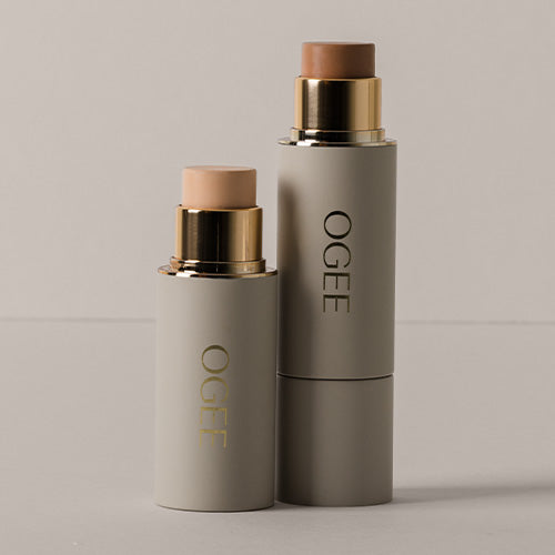 Two sage green Ogee makeup sticks stand side by side against a neutral gray background, one taller with its cap off showing a light beige formula and the other shorter.