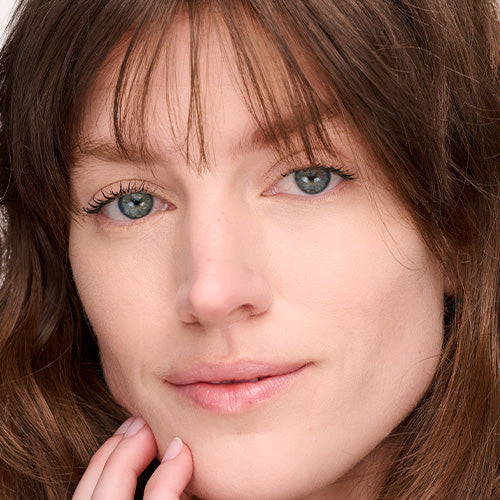 A close-up portrait of a woman with long auburn hair and bangs, light eyes, and a natural, dewy complexion, resting her fingertips against her jaw.