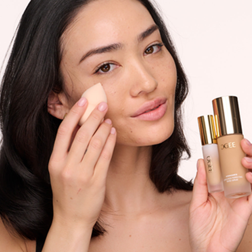 A person applies makeup to their cheek with a beauty sponge while holding two Ogee product bottles in their other hand.