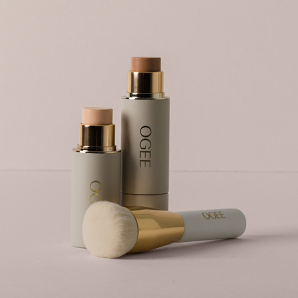 Two Ogee face sticks with gold accents, one with a tan bronzer tip and a shorter one with a pale pink tip, stand behind an Ogee makeup brush lying on a soft gray surface.