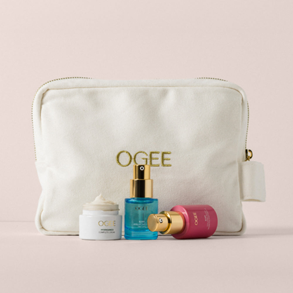 A cream-colored branded pouch embroidered “OGEE” sits behind three skincare products: an open jar of cream, a blue pump bottle, and a pink spray bottle.