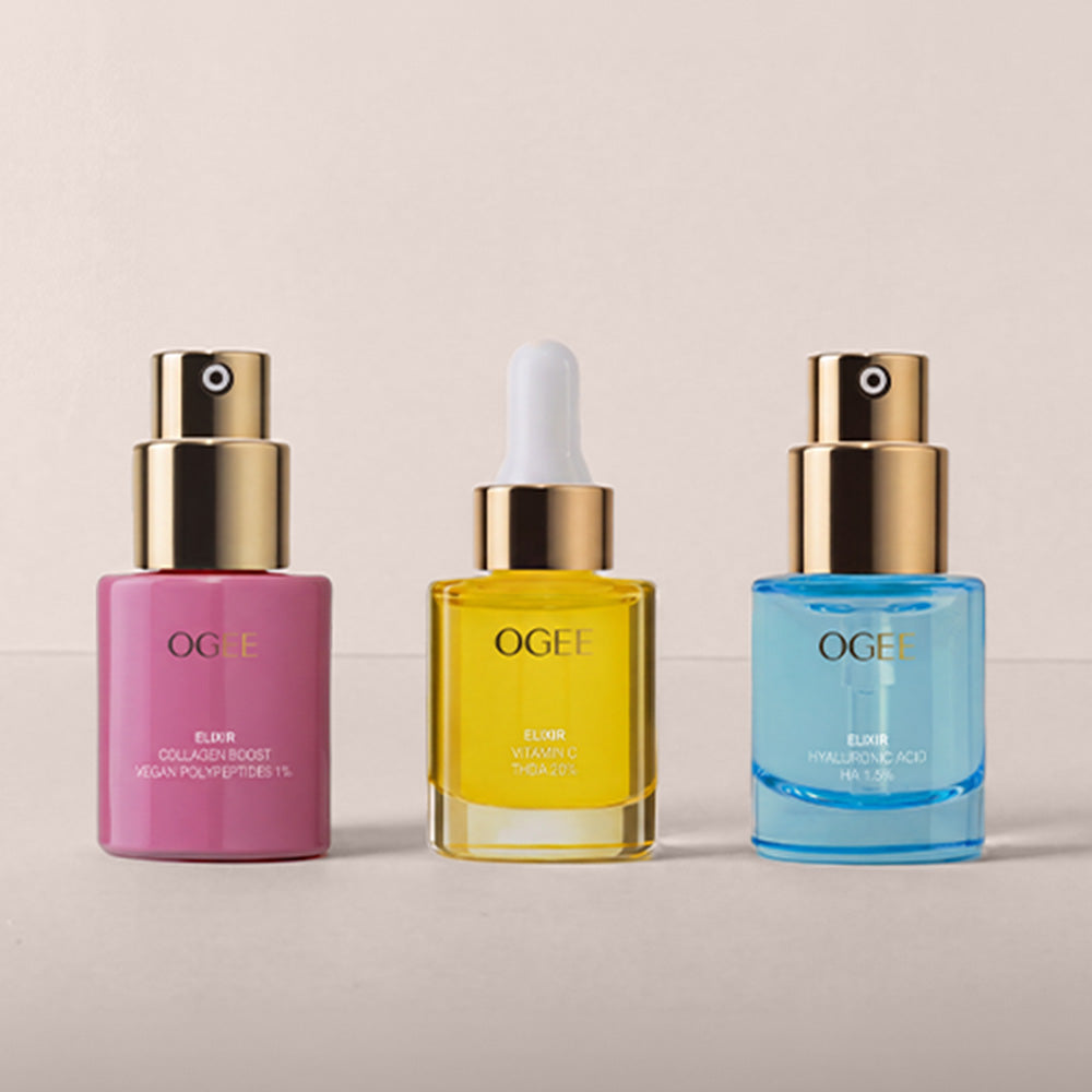 Three OGEE skincare bottles lined up against a light beige background: a pink collagen-infused serum with a gold pump, a yellow retinol dropper bottle with a white dropper top, and a blue hyaluronic acid bottle with a gold pump.