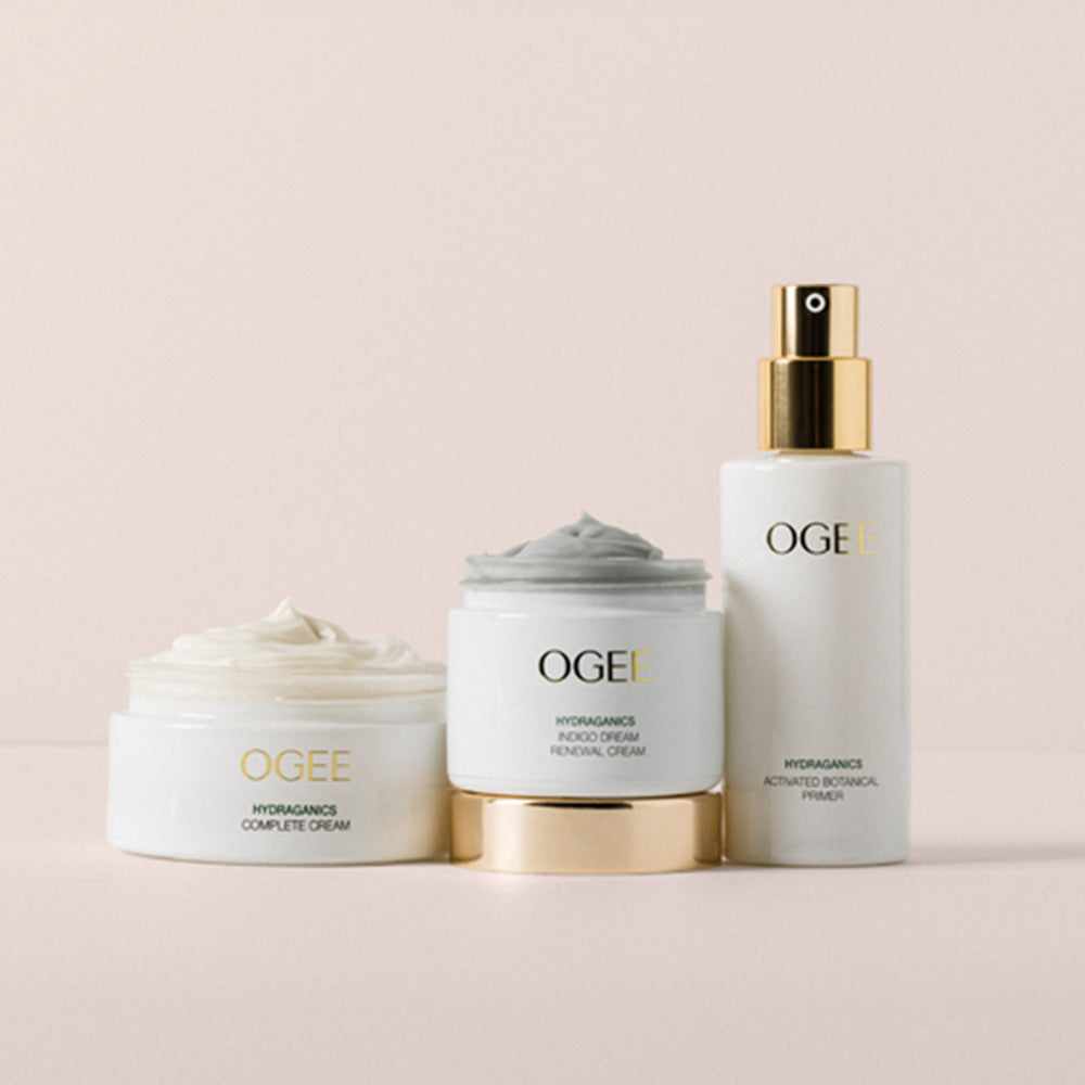 Three OGEE skincare products arranged on a soft blush background: a jar of complete cream with white cream visible, a jar of facial cream with gray cream visible and a gold base, and a tall pump bottle with a gold sprayer.