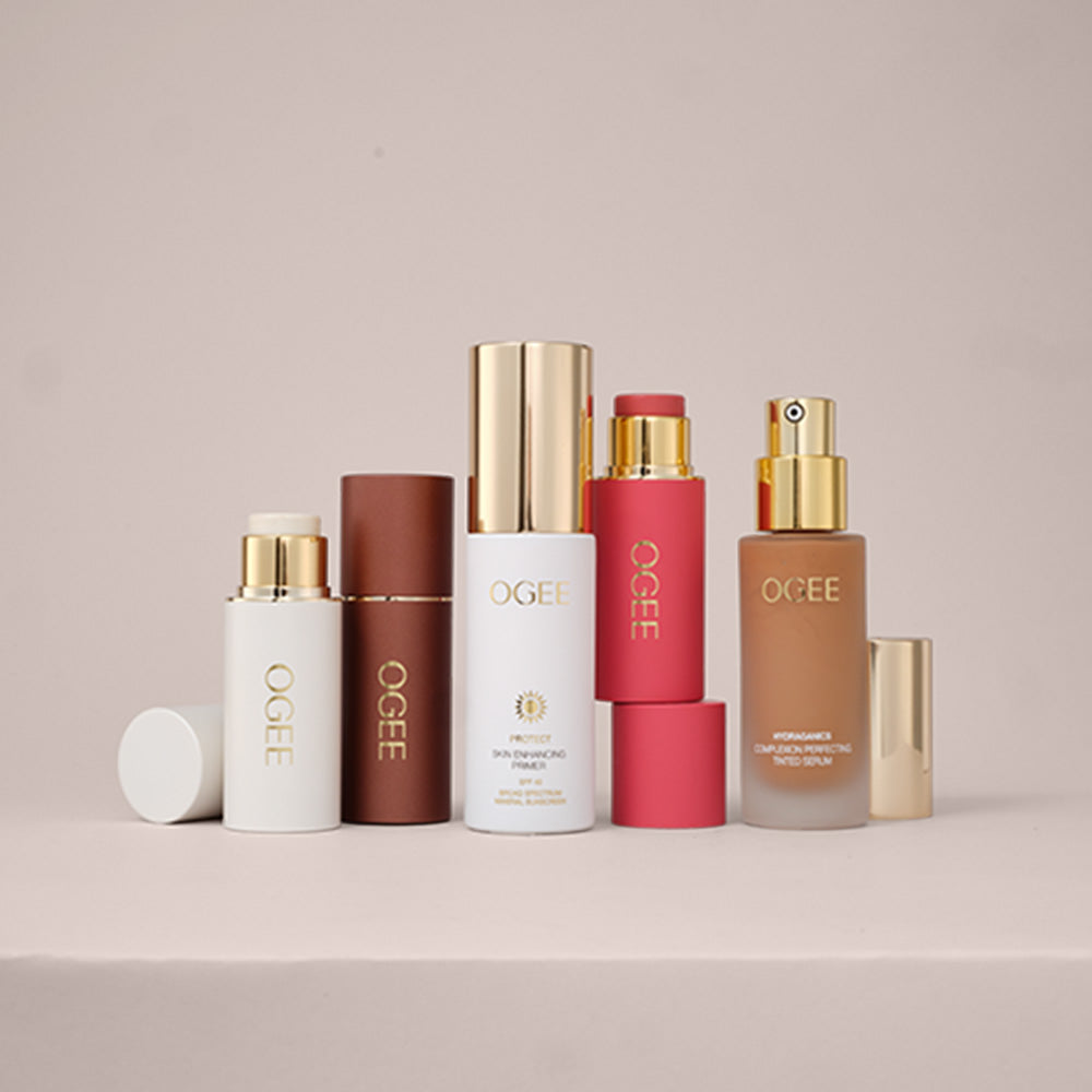 A set of OGEE cosmetic products arranged on a light beige platform against a soft beige background. The products include a white bottle with a gold cap, a brown rectangular bottle, a white bottle with a gold cap, a pink bottle with a gold cap, a pink cylindrical container, and a frosted brown bottle with a gold pump, all with gold branding.