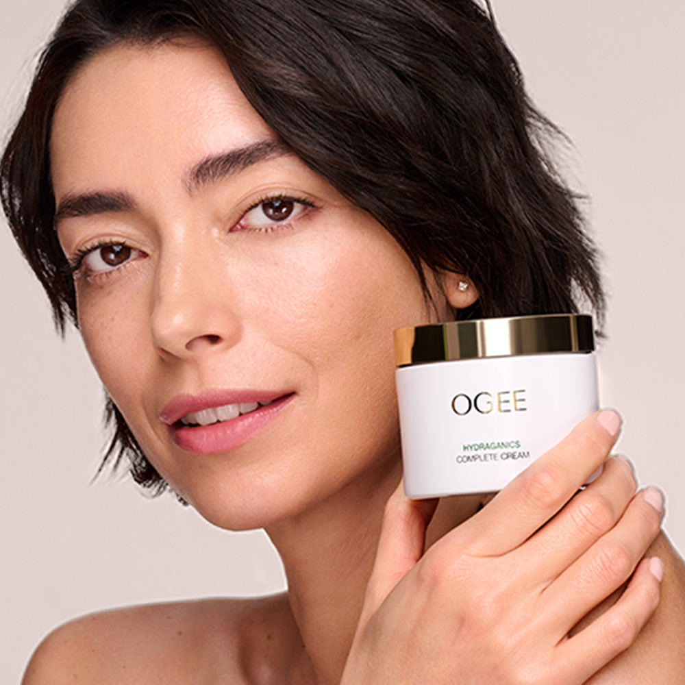 A close-up of a person’s face and shoulder as they hold a white jar labeled “OGEE HYDRAGANICS COMPLETE CREAM” near their cheek.