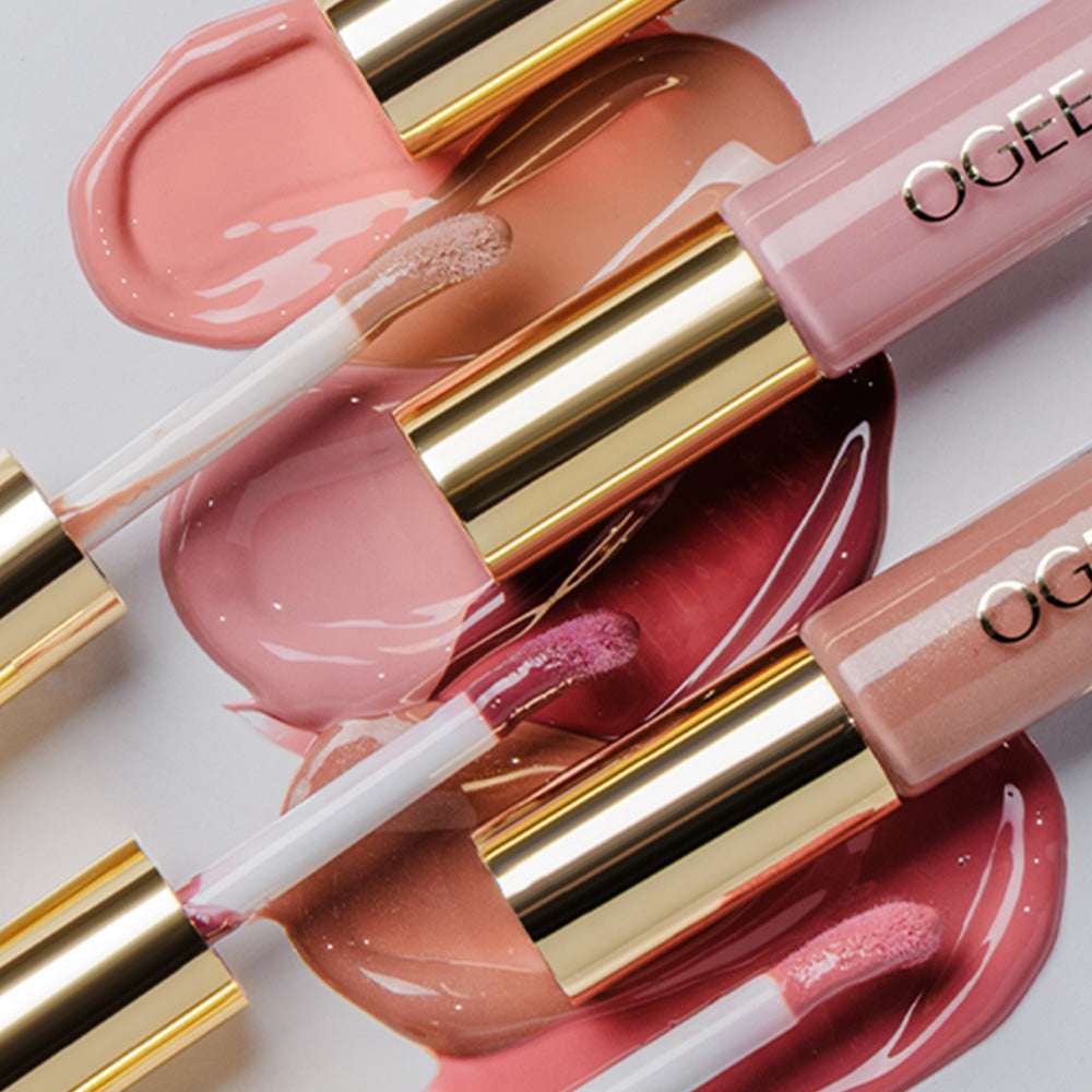 Several Ogee lip glosses in pink and nude shades with gold caps lie diagonally across glossy swatches of pink, peach, and berry gloss.